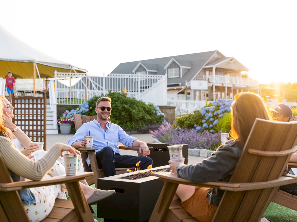 A group of friends sits around a fire pit on a sunny deck, chatting and enjoying drinks in outdoor lounge chairs. End.