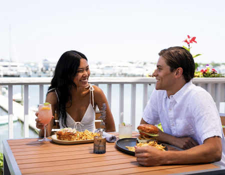 A couple sits at a sunny outdoor table by the harbor, sharing laughter over burgers and fries with drinks.