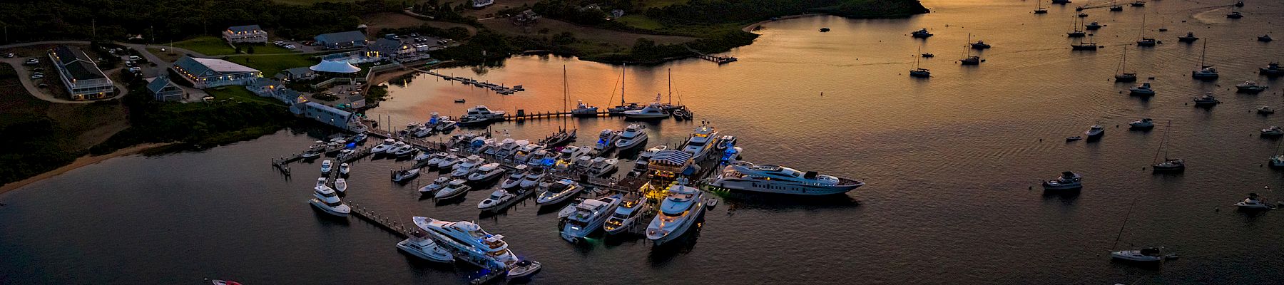 Sunset over a tranquil harbor, rows of boats clustered along the shore, calm water reflecting fiery orange and purple skies.