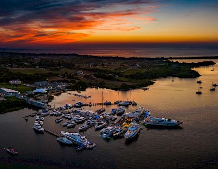 Sunset over a tranquil harbor, rows of boats clustered along the shore, calm water reflecting fiery orange and purple skies.