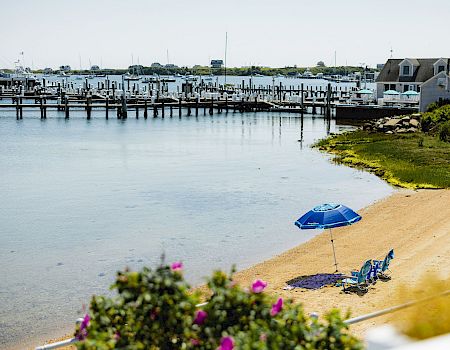 A calm harbor with a sandy beach, blue umbrella and lounge chairs by the water, a wooden pier stretching into the distance, and houses along the shore.