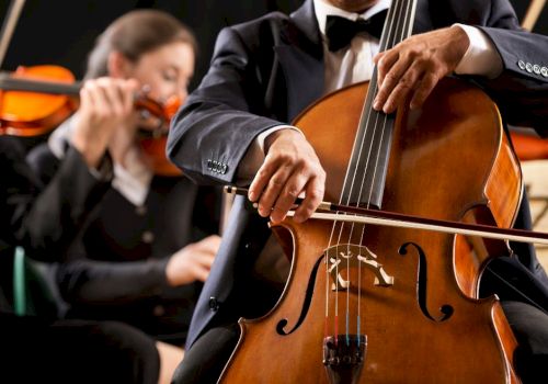 A close-up of a cellist playing a cello, with a violinist in the background during a formal orchestra performance.