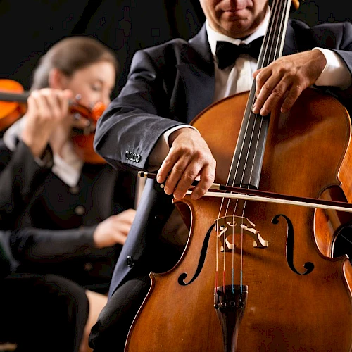 A close-up of a cellist playing a cello, with a violinist in the background during a formal orchestra performance.