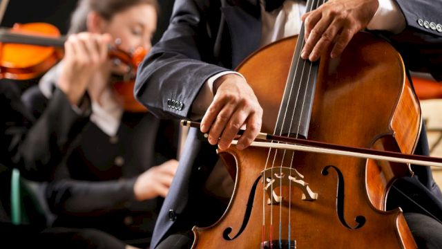 A close-up of a cellist playing a cello, with a violinist in the background during a formal orchestra performance.