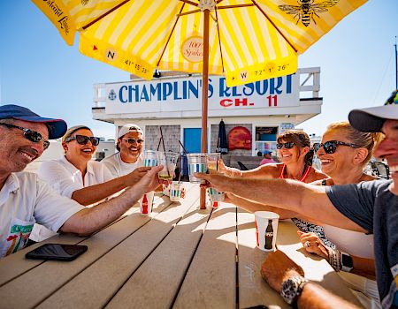 A group of people sits at a picnic table under a yellow umbrella, clinking drinks and smiling in a sunny outdoor setting near a sign that reads &ldquo;Champions&rdquo; or similar.