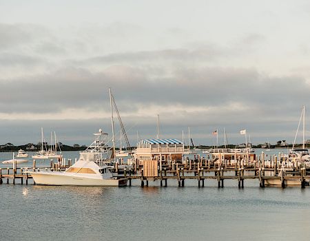 A marina with several yachts docked at wooden piers, calm water, and a cloudy sky&mdash;boats and fishing structures line the harbor, peaceful scene.
