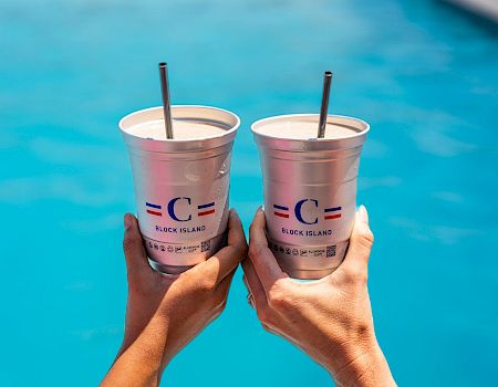 Two hands hold up two light pink cups with black straws over a blue pool, celebrating a sunny day by the water.