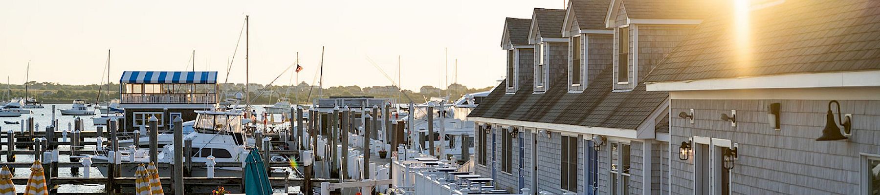 A sunny marina scene with boats docked, a row of seaside cottages, outdoor seating, and a bright sun over the harbor.