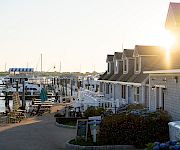 A sunny marina scene with boats docked, a row of seaside cottages, outdoor seating, and a bright sun over the harbor.