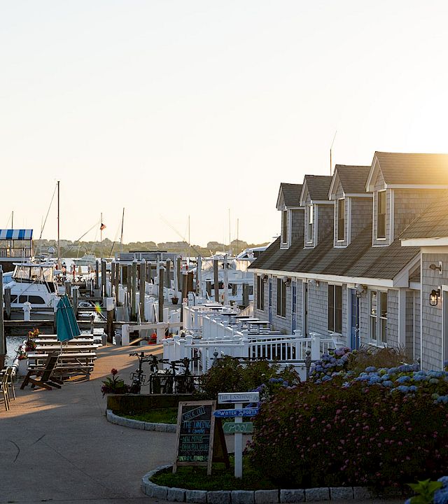 A sunny marina scene with boats docked, a row of seaside cottages, outdoor seating, and a bright sun over the harbor.