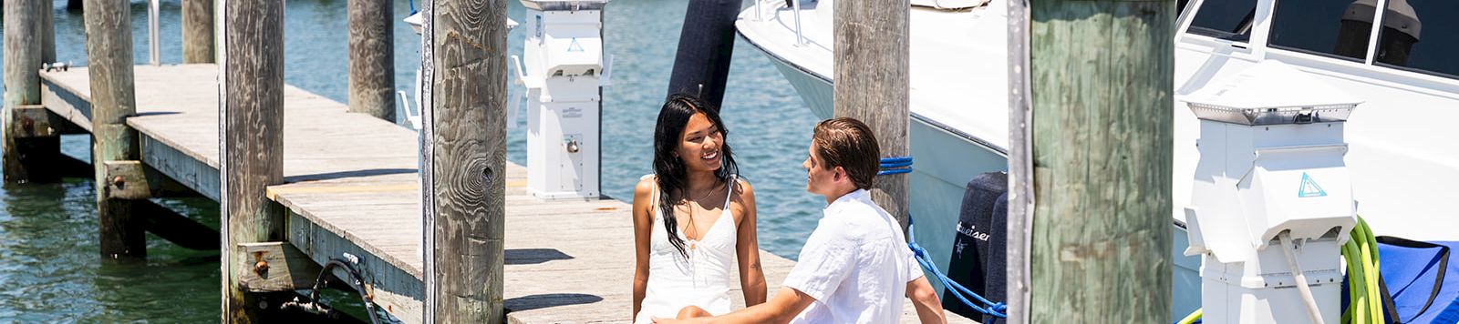 Two people sit on a wooden dock by the water, chatting with boats and masts in the background on a sunny day.