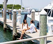 Two people sit on a wooden dock by the water, chatting with boats and masts in the background on a sunny day.