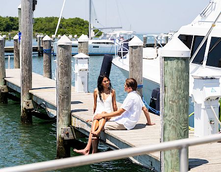Two people sit on a wooden dock by the water, chatting with boats and masts in the background on a sunny day.