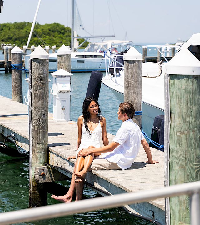 Two people sit on a wooden dock by the water, chatting with boats and masts in the background on a sunny day.