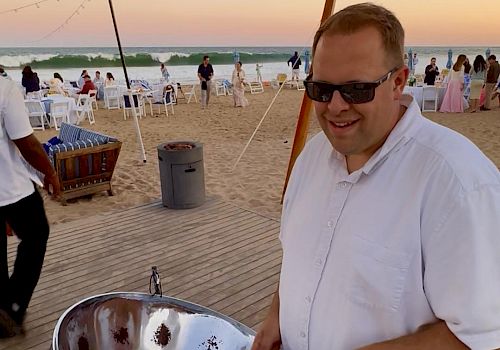 A smiling man in a white shirt wearing sunglasses grilling on a shiny metal bowl at a beach party, with sunset, string lights, and people in the background, all on a wooden deck.