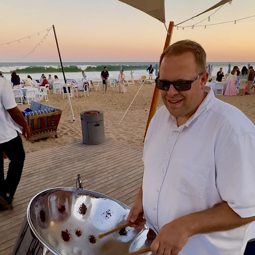 A smiling man in a white shirt wearing sunglasses grilling on a shiny metal bowl at a beach party, with sunset, string lights, and people in the background, all on a wooden deck.