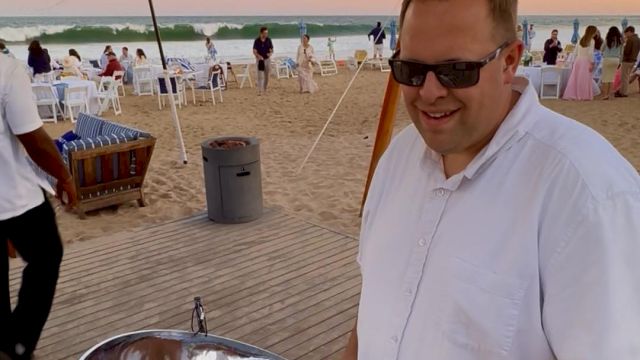 A smiling man in a white shirt wearing sunglasses grilling on a shiny metal bowl at a beach party, with sunset, string lights, and people in the background, all on a wooden deck.