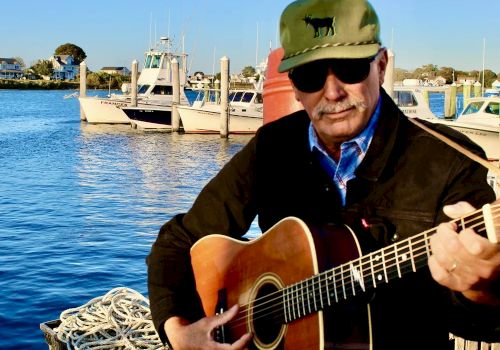 A man with a guitar by the water, wearing a green cap and sunglasses, playing beside boats and a dock at sunset, surfacing calm vibes.