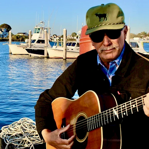 A man with a guitar by the water, wearing a green cap and sunglasses, playing beside boats and a dock at sunset, surfacing calm vibes.