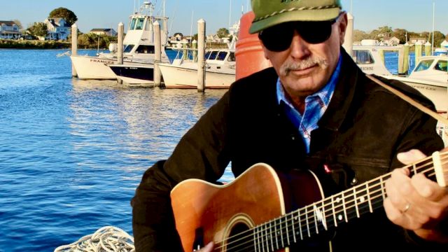 A man with a guitar by the water, wearing a green cap and sunglasses, playing beside boats and a dock at sunset, surfacing calm vibes.
