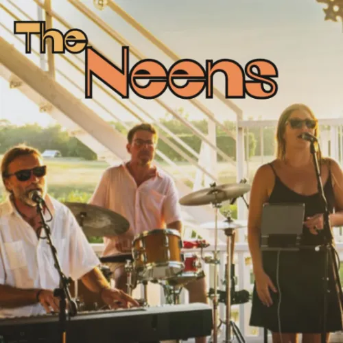 The Neens live band performs on stage with two men on percussion and guitar and a singer with a microphone and sunglasses, outdoors at sunset.