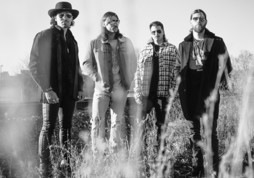 A black-and-white photo of four men standing in tall grass outdoors, wearing casual jackets and hats, posing for a band-style portrait.