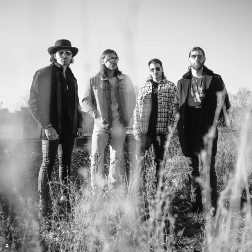 A black-and-white photo of four men standing in tall grass outdoors, wearing casual jackets and hats, posing for a band-style portrait.