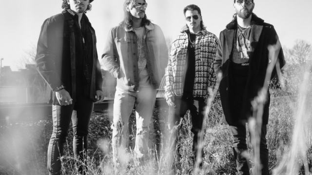A black-and-white photo of four men standing in tall grass outdoors, wearing casual jackets and hats, posing for a band-style portrait.