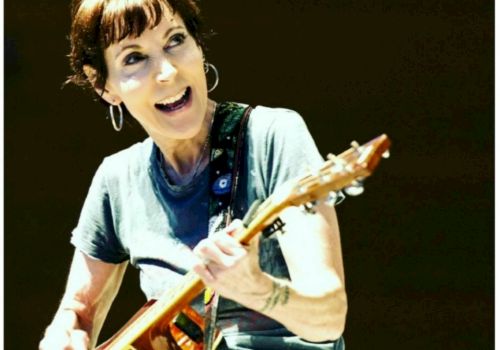 Carrie Johnson performing with an acoustic guitar, smiling while playing on stage. Looking up, energetic live moment.