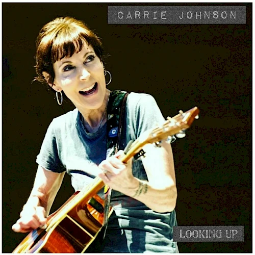 Carrie Johnson performing with an acoustic guitar, smiling while playing on stage. Looking up, energetic live moment.