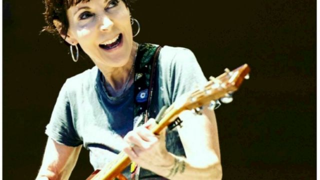 Carrie Johnson performing with an acoustic guitar, smiling while playing on stage. Looking up, energetic live moment.