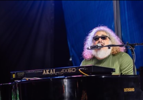 A bearded man with sunglasses plays a Yamaha Akai AX60 keyboard on a stage, singing into a microphone under blue lighting.