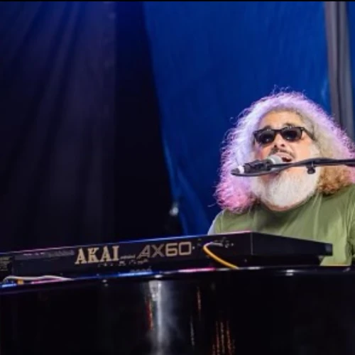 A bearded man with sunglasses plays a Yamaha Akai AX60 keyboard on a stage, singing into a microphone under blue lighting.