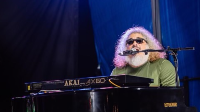 A bearded man with sunglasses plays a Yamaha Akai AX60 keyboard on a stage, singing into a microphone under blue lighting.