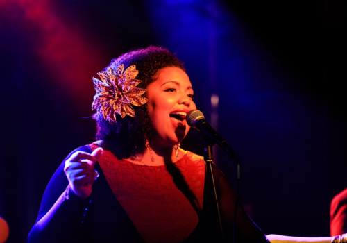 A joyful female singer with a vintage flower in her hair performs into a microphone on a moody, colorful stage, glowing with red and blue lights.