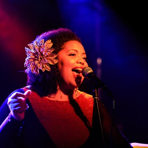A joyful female singer with a vintage flower in her hair performs into a microphone on a moody, colorful stage, glowing with red and blue lights.