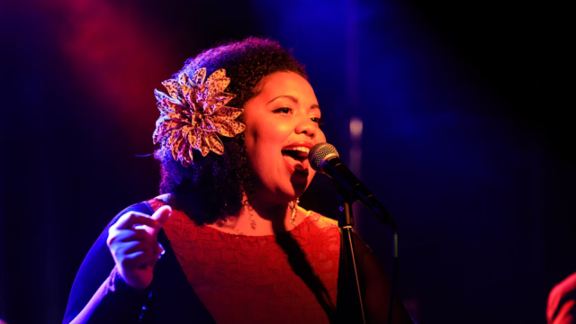 A joyful female singer with a vintage flower in her hair performs into a microphone on a moody, colorful stage, glowing with red and blue lights.