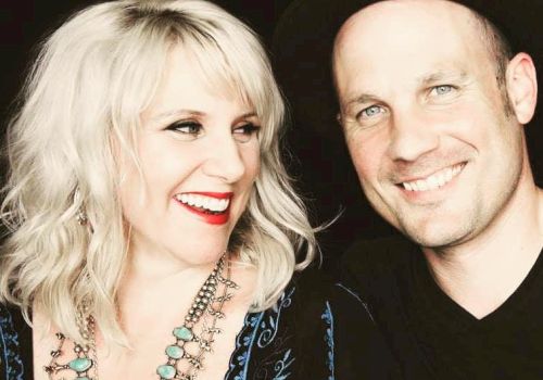 A smiling blonde woman with turquoise necklace and red lipstick sits beside a man in a black hat; both look cheerful and pose for a photo.