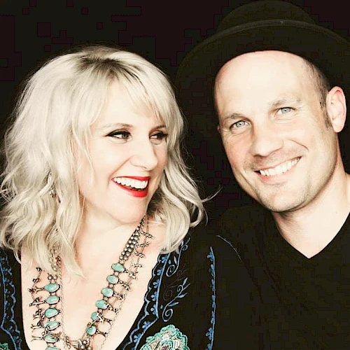 A smiling blonde woman with turquoise necklace and red lipstick sits beside a man in a black hat; both look cheerful and pose for a photo.