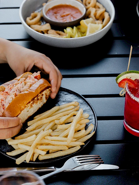 A person is eating a burger and fries at an outdoor table, with a red cocktail nearby and another dish in the background. End with a period.