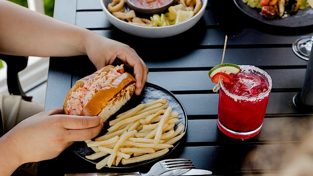 A person is eating a burger and fries at an outdoor table, with a red cocktail nearby and another dish in the background. End with a period.