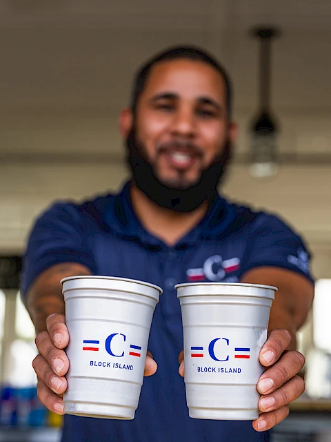 Two cups held toward the camera by a smiling man in a blue polo, serving drinks in a bright indoor setting.