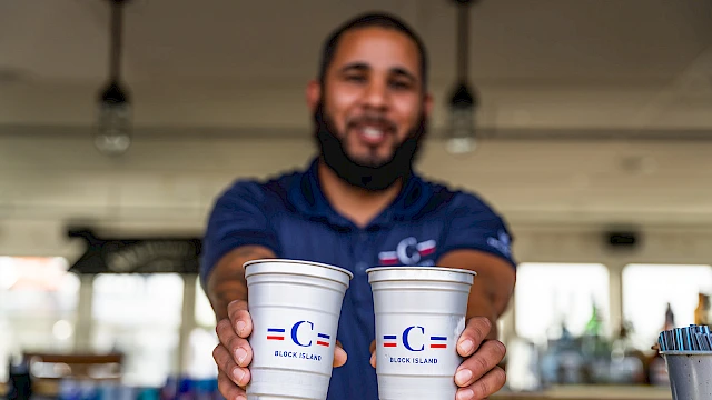 Two cups held toward the camera by a smiling man in a blue polo, serving drinks in a bright indoor setting.