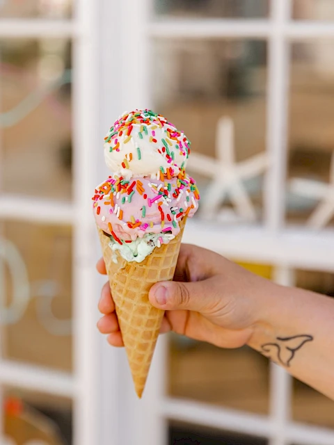 A hand holds a two-scoop cone with colorful sprinkles, set against a bright windowed background, summer treat vibe.