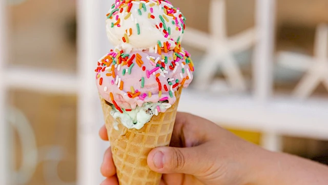 A hand holds a two-scoop cone with colorful sprinkles, set against a bright windowed background, summer treat vibe.