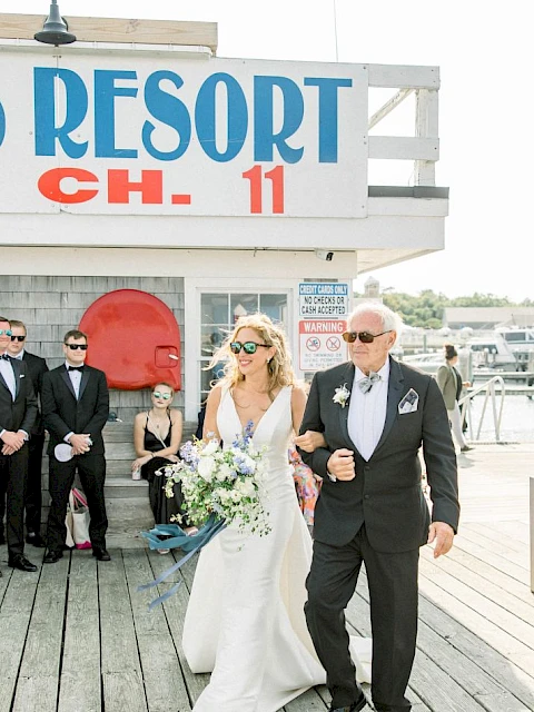 A bride in a white gown and a suited man walk arm-in-arm down a wooden boardwalk, wedding guests in masks watch beside a blue-and-white resort sign.