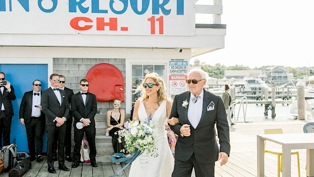 A bride in a white gown and a suited man walk arm-in-arm down a wooden boardwalk, wedding guests in masks watch beside a blue-and-white resort sign.