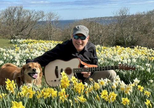 A man with a guitar sits among a field of yellow daffodils with a happy dog, scenic blue sky and distant trees in bloom.