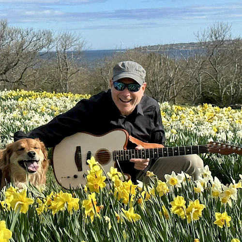 A man with a guitar sits among a field of yellow daffodils with a happy dog, scenic blue sky and distant trees in bloom.