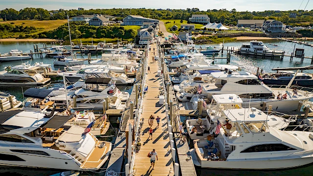 A bustling marina with boats docked on both sides of a long wooden pier, people strolling along, bright blue sky, and calm waterfront.